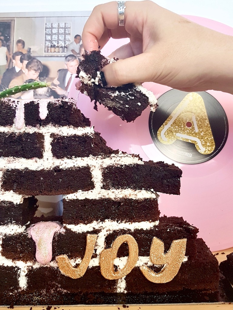 Hand grabbing a slice of beer chocolate cake built as a wall next to Idles Joy as an act of resistance vinyl record